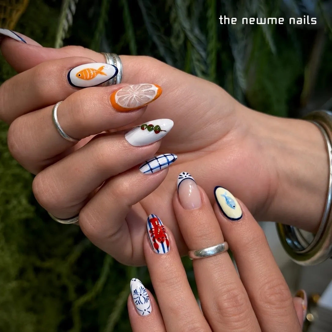 Almond nails each painted with a different motif: an orange fish, a lemon slice, olives, a blue gingham check, a red lobster on stripes and blue-and-white tile patterns.