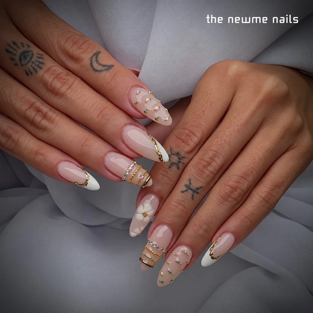 Almond nails on a sheer pink base with white french tips edged in gold, gold rings wrapped around two nails, scattered tiny gems and one painted white flower.