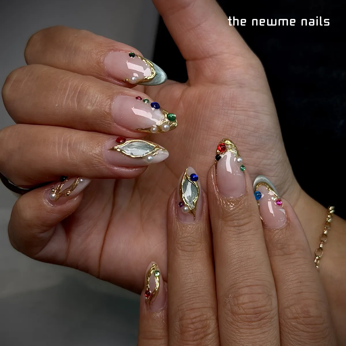 Almond nails on a sheer nude base, each framed in raised gold with small coloured gems, pearls and a pale green marbled stone.