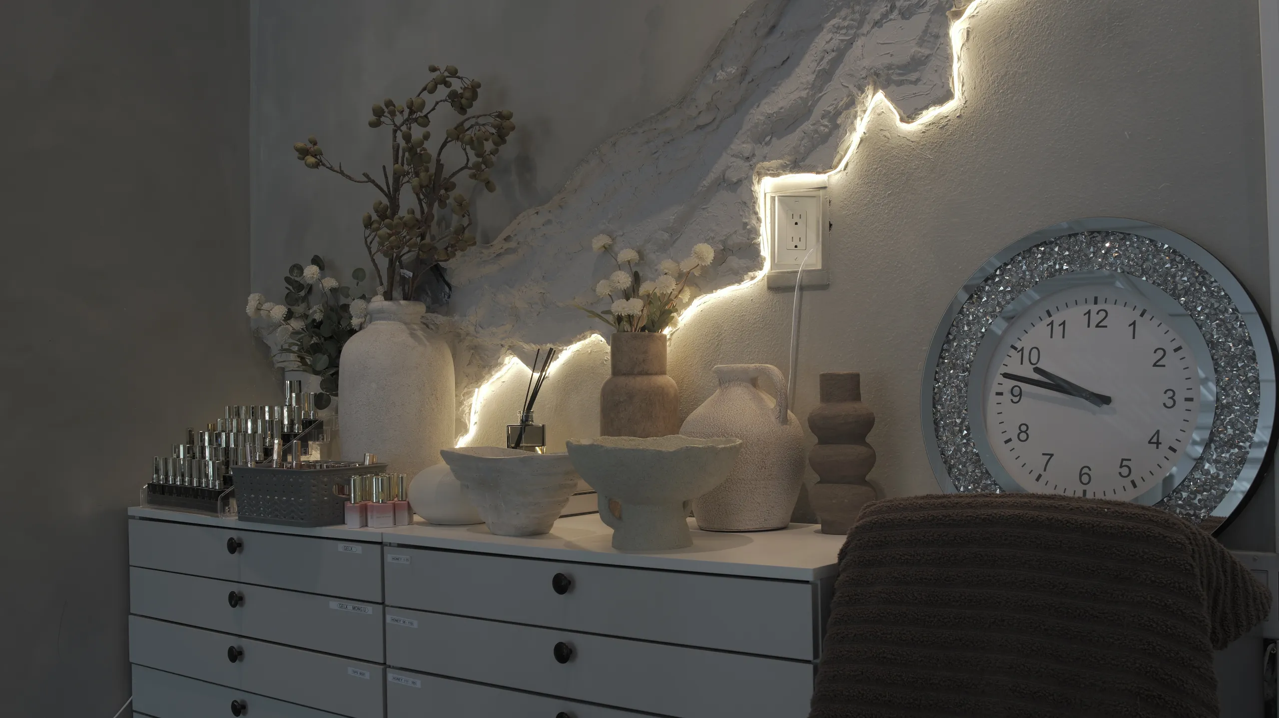 Pale ceramic vases, dried flowers and racks of polish on a white cabinet, lit by a strip of warm light along a textured plaster wall.
